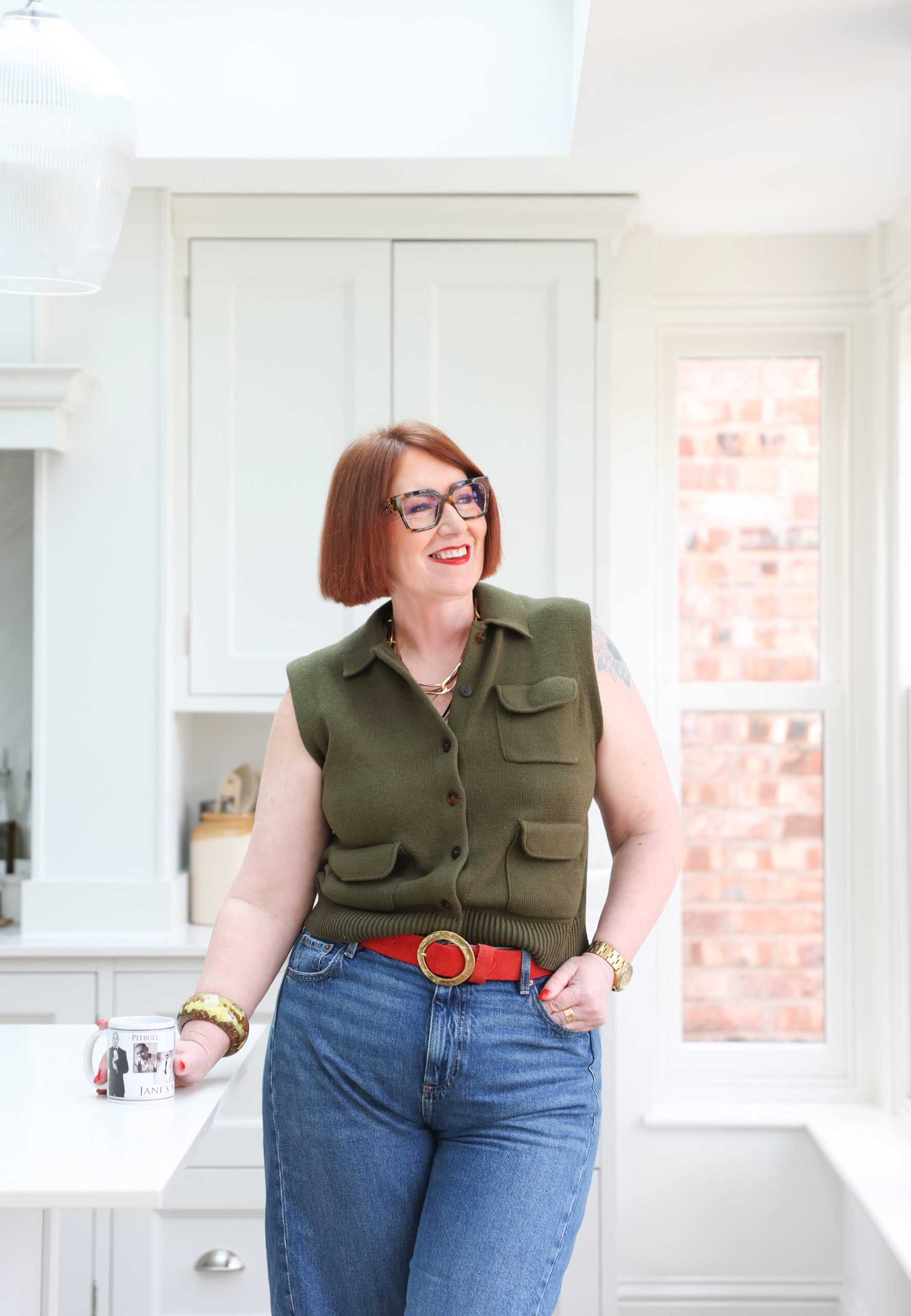 Dress for the Woman You’re Becoming Jane Brook standing in the kitchen wearing a khaki top, blue jeans and a red belt.